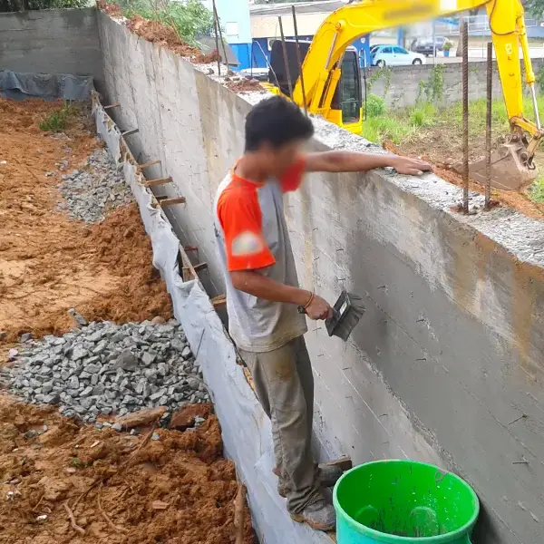 Operário fazendo o acabamento do topo do muro, com brita e tubos de drenagem ao lado